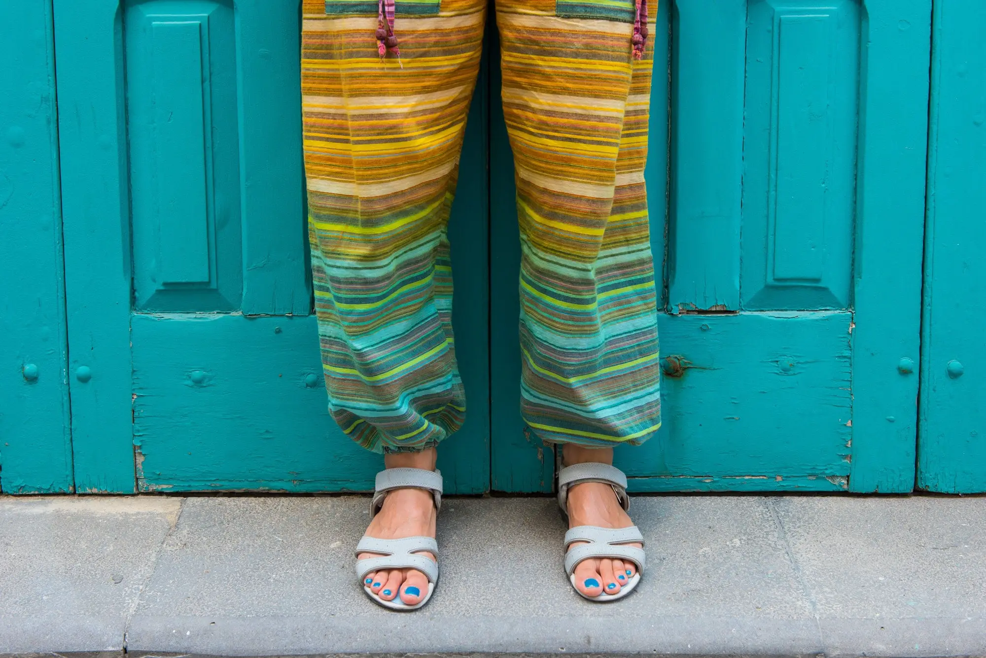 Feet of a woman in sandals with blue toe nails. Colorful hippie pants. Detail