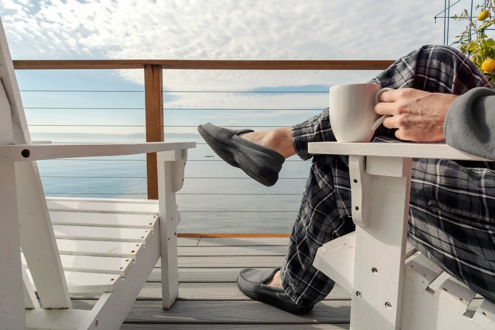 Person sitting on seaside deck wearing slippers drinking coffee.