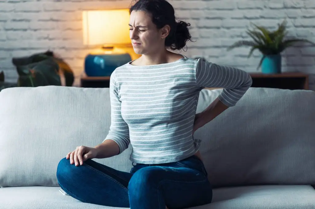 Young woman with back pain sitting on the sofa in the living room at home.