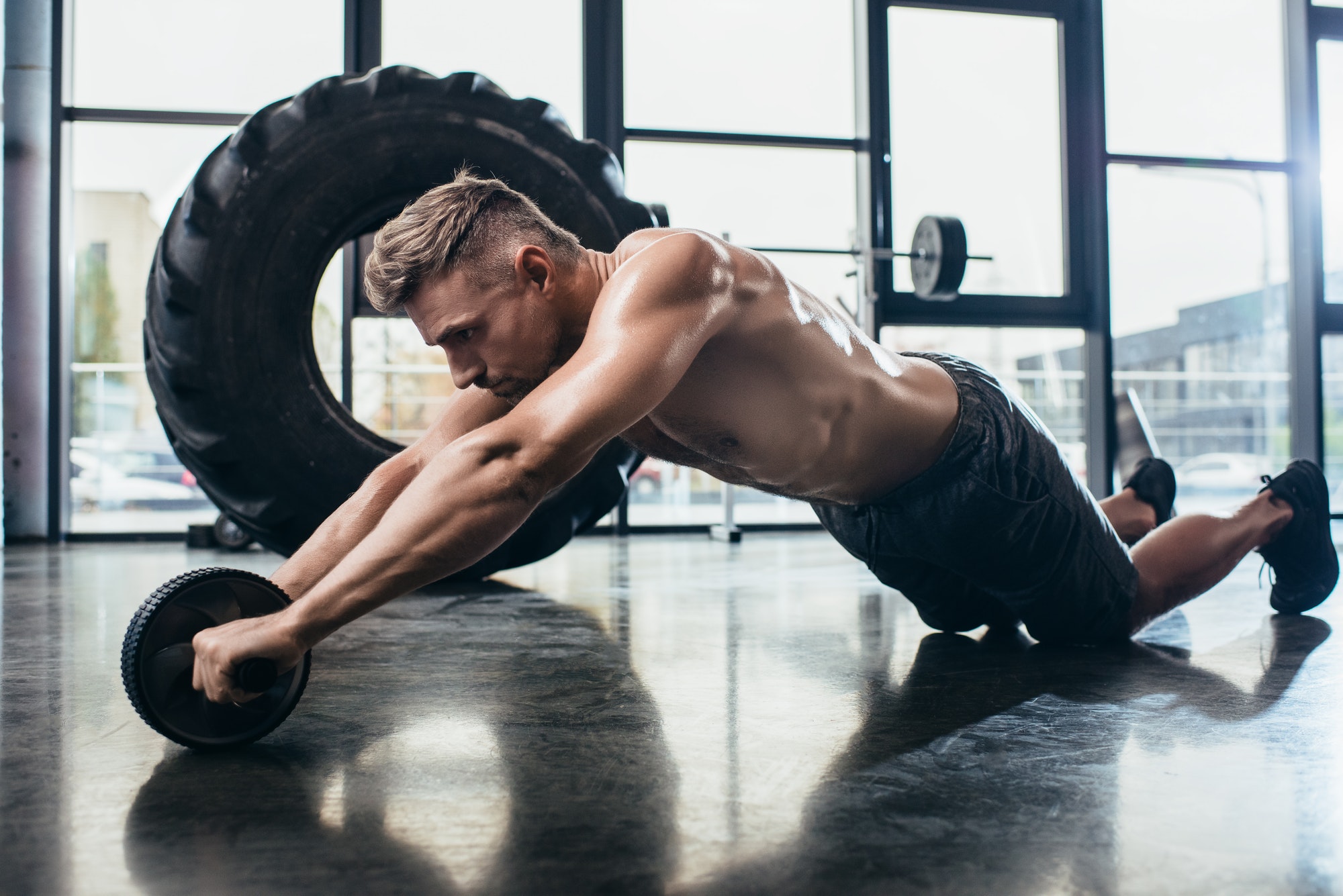 handsome shirtless muscular sportsman exercising with abs roller in gym