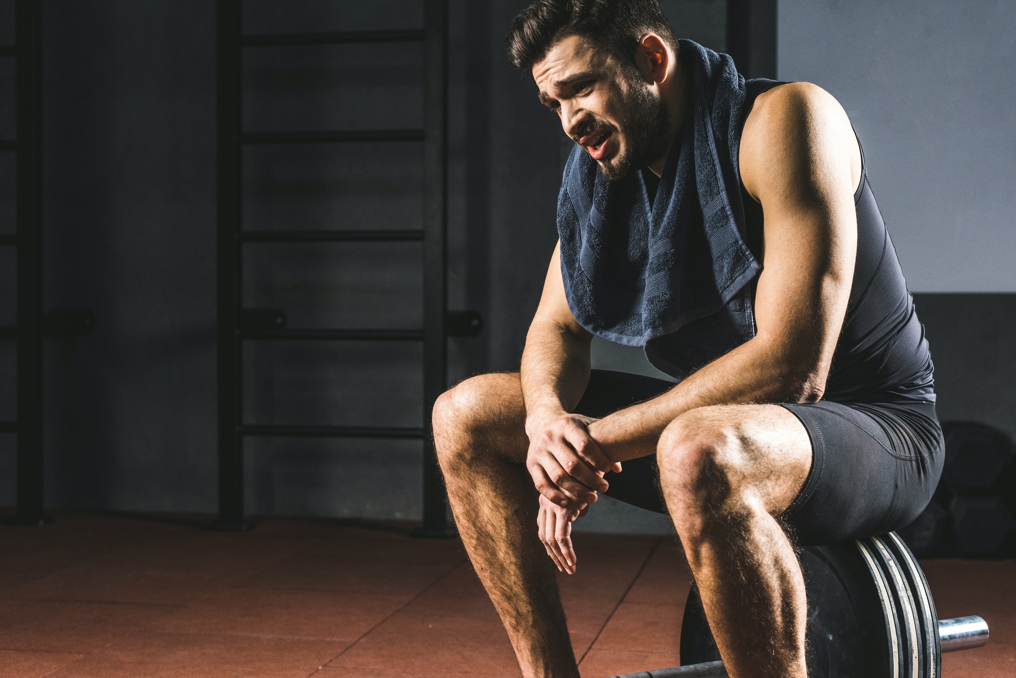 Tired young sportsman with towel on neck sitting on barbell in gym